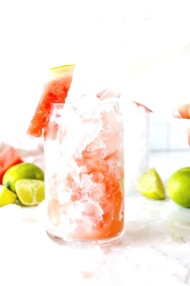 This is a side view of a watermelon cocktail being poured into a glass with crushed ice. The glass sits on a white counter with more ice and limes all around it.