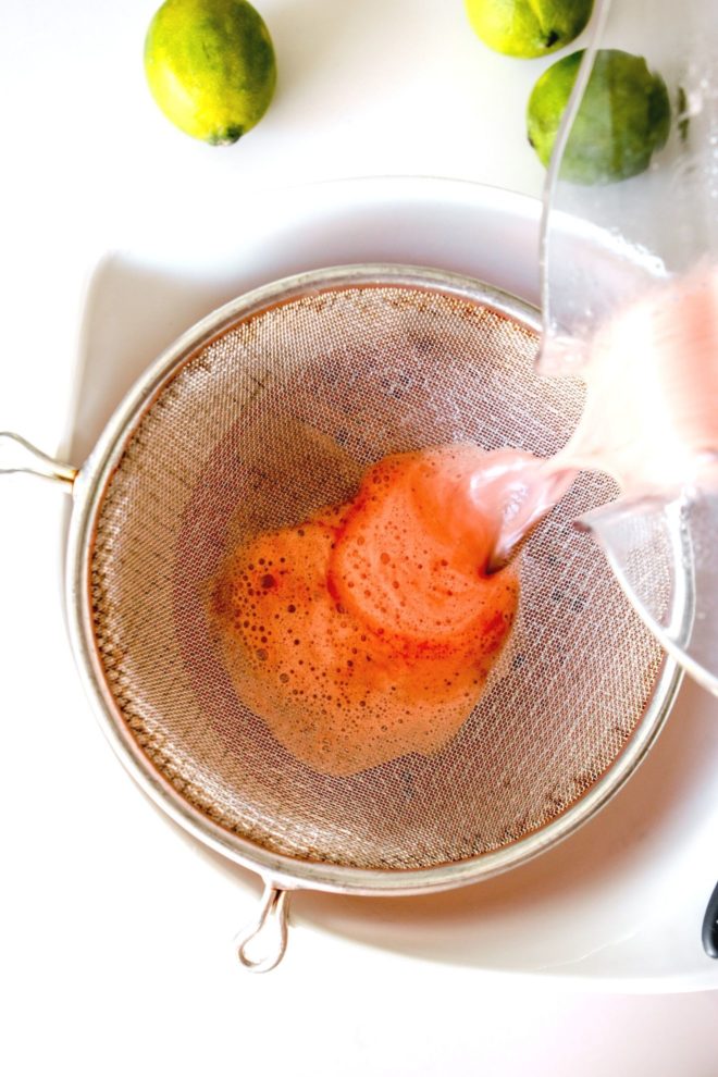 This is an overhead image of watermelon juice being poured through a strainer into a white bowl. The bowl sits on a white counter with limes to the top of the image.
