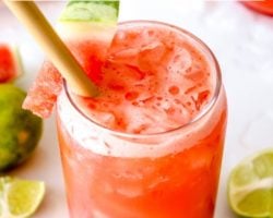 This is a side view looking into the top of a glass filled with a watermelon cocktail. The glass is garnished with a watermelon spear. The glass sits on a white counter with melted ice and more watermelon and lime pieces around it. Another full glass is in the top right of the image and a light pink towel is in the top left of the image. Text overlay reads "easy & delicious watermelon vodka cocktail"