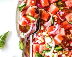 This is an overhead image of a watermelon salad with red onion, feta, pine nuts, and cucumber. The salad is on a white platter on a white counter with basil leaves around the platter. Two large silver spoons are scooping up some salad and laying on the side of the platter.
