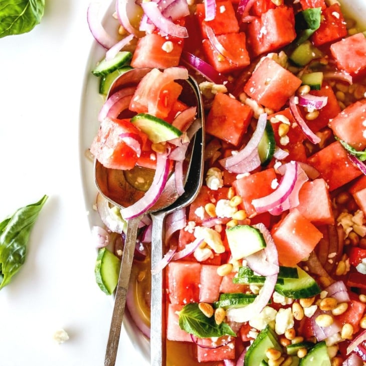 This is an overhead image of a watermelon salad with red onion, feta, pine nuts, and cucumber. The salad is on a white platter on a white counter with basil leaves around the platter. Two large silver spoons are scooping up some salad and laying on the side of the platter.