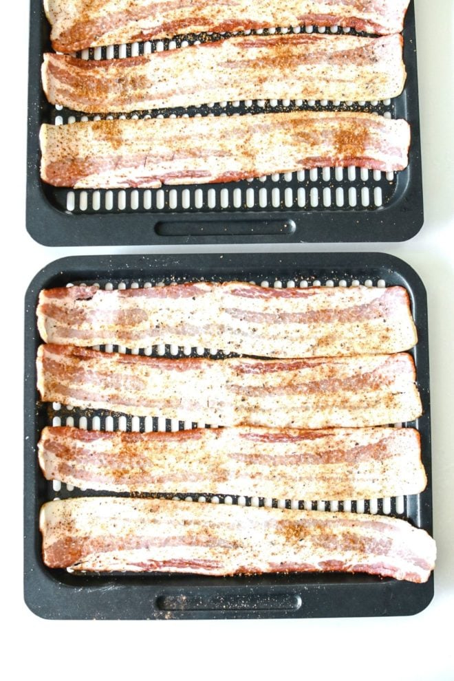 This is an overhead image of slices of bacon on an air fryer tray. The raw bacon slices are sprinkled with sugar and pepper. The trays are on a white counter.