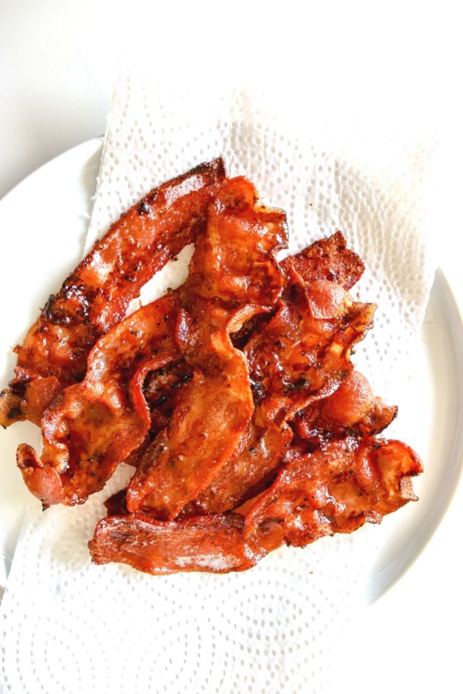 This is an overhead image of cooked slices of bacon on a white plate lined with a paper towel. The plate sits on a white counter.