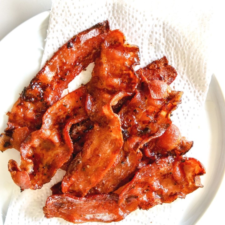 This is an overhead image of cooked slices of bacon on a white plate lined with a paper towel. The plate sits on a white counter.