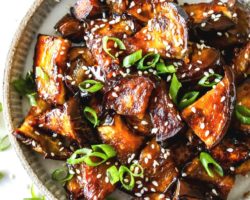 This is an overhead image of a plate with caramelized, glazed eggplant pieces. The eggplant has sesame seeds and scallions on it. The plate sits on a white counter.