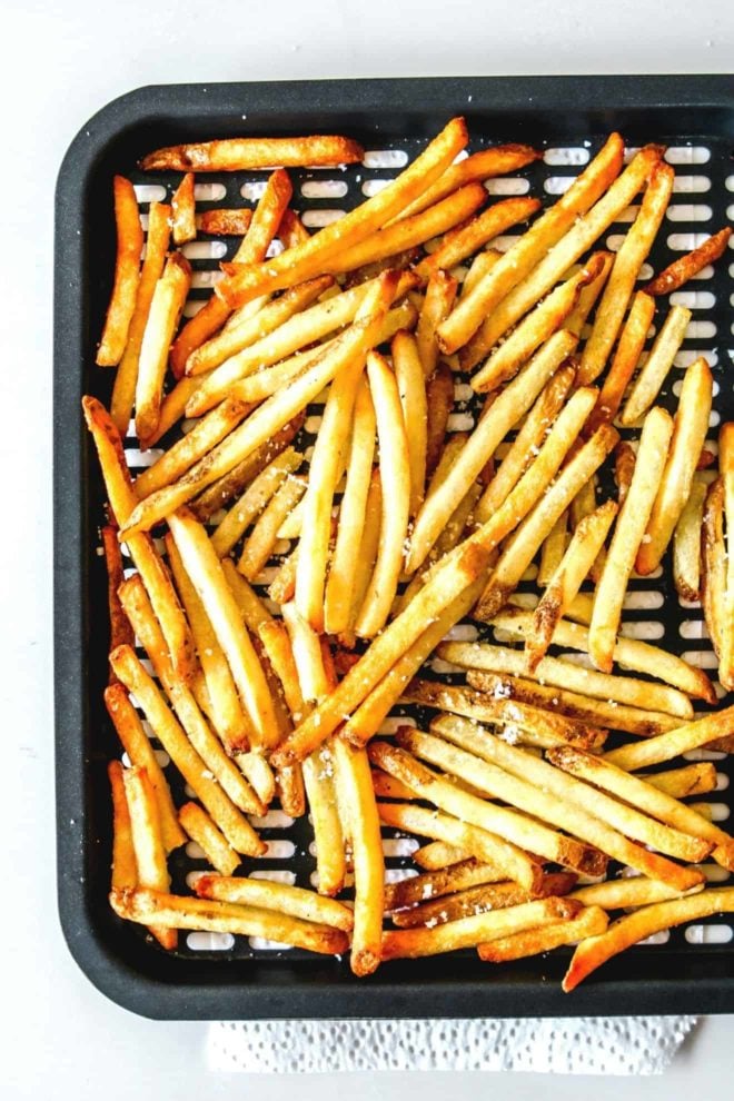 This is an overhead image of an air fryer tray with cooked french fries on it. The fries are sprinkled with salt. The tray is on a white paper towel and on a white counter.