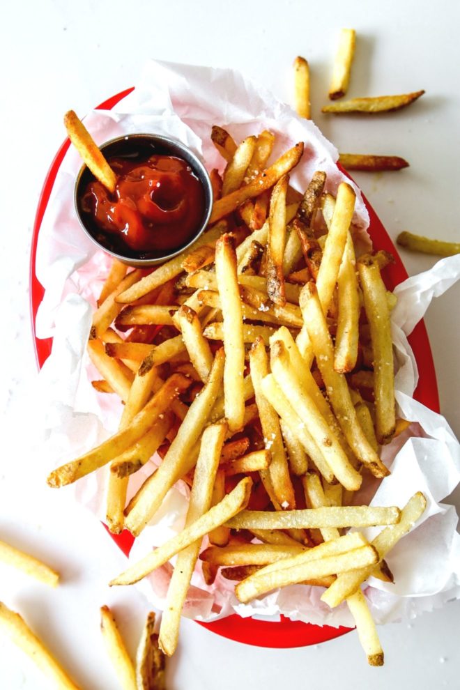 This is an overhead image of a red plastic basket with white parchment paper and french fries sprinkled with salt. One french fry dipping into a small metal bowl of ketchup. The basket sits on a white counter.