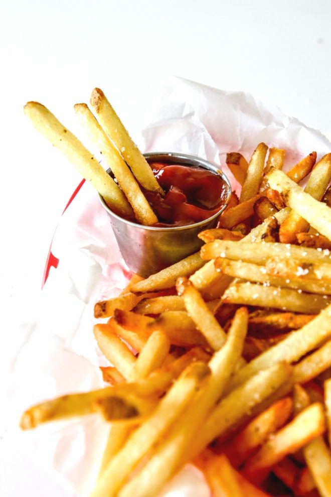 This is a side view of a red plastic basket with white parchment paper and french fries sprinkled with salt. Three french fries are dipping into a small metal bowl of ketchup. The basket sits on a white counter.