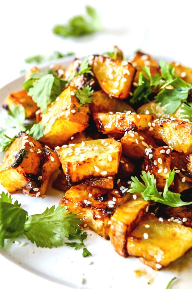 This is a side view of a white plate on a white counter. On the plate are pieces of glazed sweet potato topped with sesame seeds and cilantro.