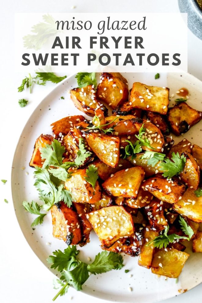 This is an overhead view of a white plate on a white counter. On the plate are pieces of glazed sweet potato topped with sesame seeds and cilantro. To the top right corner is a small blue bowl with white sesame seeds in it. Text overlay reads "miso glazed air fryer sweet potatoes."