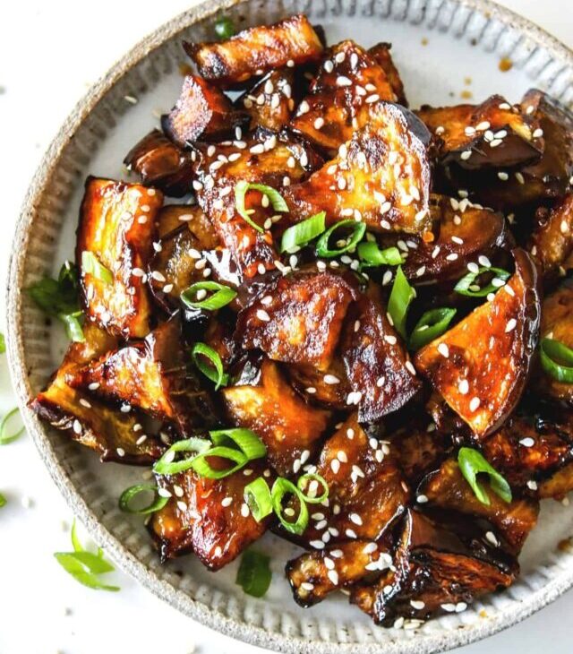 This is an overhead image of a plate with caramelized, glazed eggplant pieces. The eggplant has sesame seeds and scallions on it. The plate sits on a white counter.