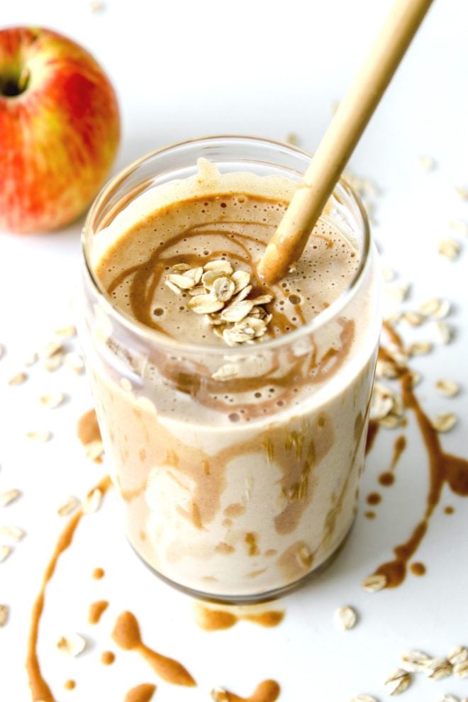 This is view looking into a glass with a smoothie and some peanut butter dripping inside the glass and on top of the smoothie. The glass is sitting on a white counter with some peanut butter and oats around it. There is an apple blurred in the top left of the image.
