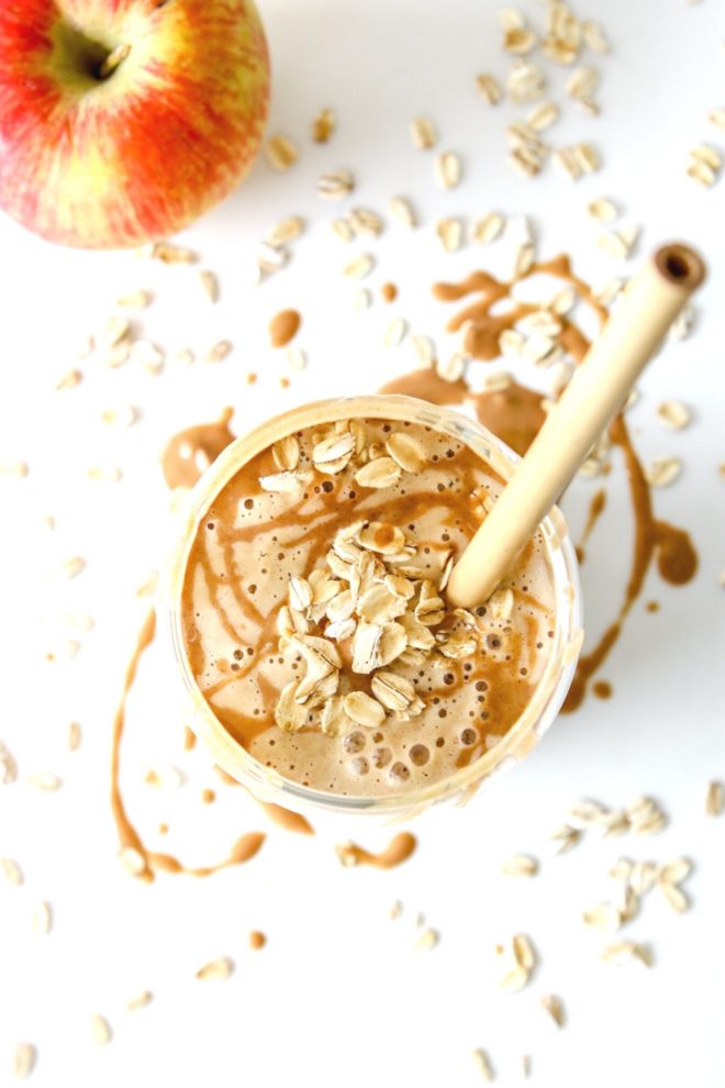 This is an overhead image of a glass with a tan smoothie in it. The glass has peanut butter and oats on top and a straw sticking out. The glass sits on a white counter with peanut butter and oats around it. An apple is in the top left corner of the image.