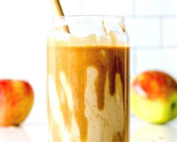 This is a side view of a glass with a smoothie and some peanut butter dripping inside the glass. The glass is sitting on a white counter with some peanut butter and oats. There are two apples blurred in the background.