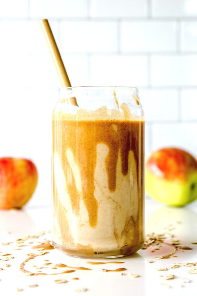This is a side view of a glass with a smoothie and some peanut butter dripping inside the glass. The glass is sitting on a white counter with some peanut butter and oats. There are two apples blurred in the background.