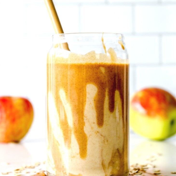 This is a side view of a glass with a smoothie and some peanut butter dripping inside the glass. The glass is sitting on a white counter with some peanut butter and oats. There are two apples blurred in the background.