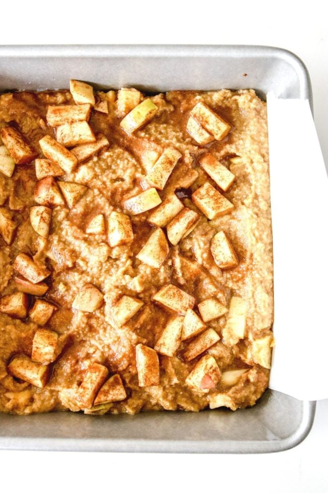This is an overhead image of a square pan with raw blondie batter. The blondies are topped with raw cinnamon coated apple pieces. The pan sits on a white counter.