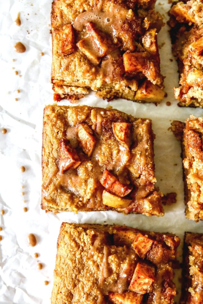 This is an overhead image of apple blondies cut into squares. The image focuses on three blondies with more to the righthand side of the image. On top of the blondies are pieces of apple and a drizzle of cinnamon icing. More icing droplets are on the parchment paper to the left of the blondies.