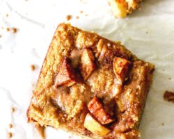 This is an overhead image of an apple blondie on a white piece of parchment paper. The blondie is drizzled with a cinnamon icing. Another blondie is to the top right corner of the image.