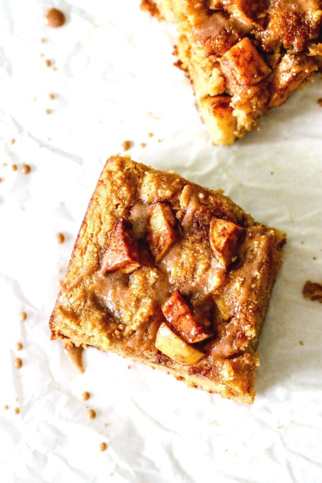 This is an overhead image of an apple blondie on a white piece of parchment paper. The blondie is drizzled with a cinnamon icing. Another blondie is to the top right corner of the image.