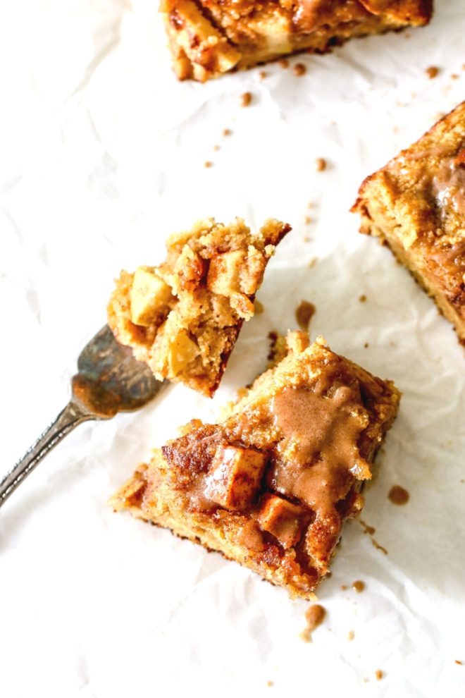 This is an overhead image of an apple blondie on a white piece of parchment paper. A fork has a bite of the blondie and is laying on counter next to the blondie. More blondies are to the top and right of the image.
