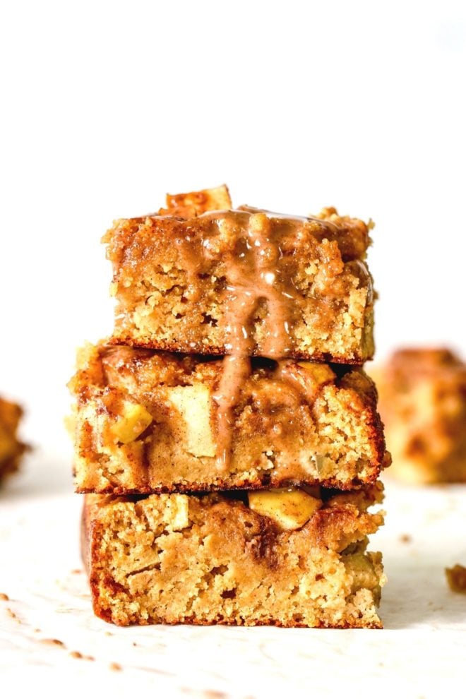 This is a stack of three apple blondies sitting on a white counter. A cinnamon icing is drizzling down the side of the stack. More apple blondies are blurred in the background.
