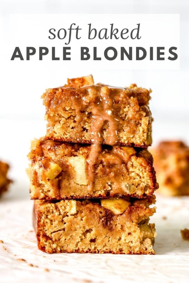 This is a stack of three apple blondies sitting on a white counter. A cinnamon icing is drizzling down the side of the stack. More apple blondies are blurred in the background. Text overlay reads "soft baked apple blondies."