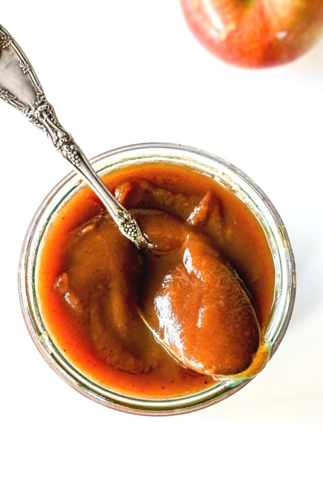 This is an overhead image of a glass jar with apple butter. An antique spoon is scooping some apple butter and leaning against the top of the jar. The glass is on a white counter and an apple is in the top right corner of the image.