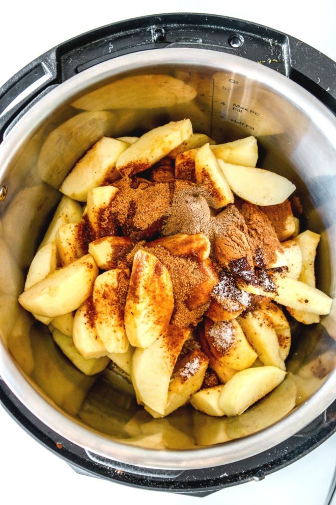This is an overhead image looking into an instant pot. In the instant pot are cut apples, coconut sugar, cinnamon, and spices. The instant pot sits on a white counter.