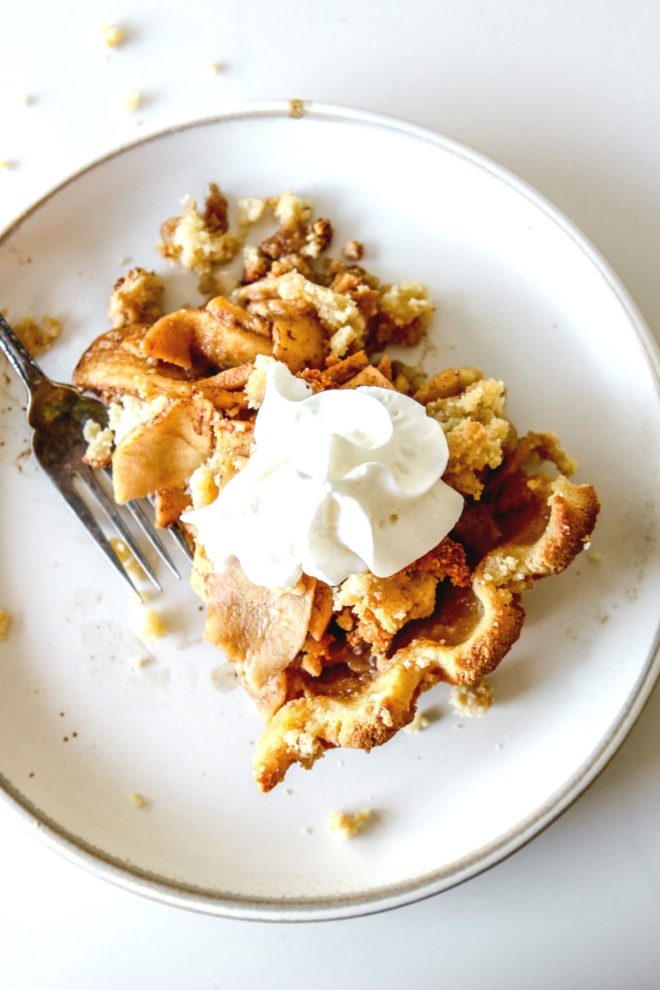 This is an overhead image of a plate with a slice of apple pie. The pie has a dollop of whipped scream on top. The plate is on a white counter and a fork is laying on the left side of the plate.