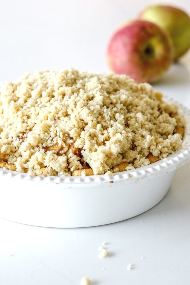 This is a side view of a white pie dish with apple pie with a crumble on top. The pie dish sits on a white counter with two apples blurred in the background.