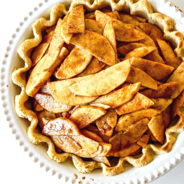 This is an overhead image of an apple pie in a white pie dish with a cooked pie crust and raw apples coated with cinnamon in it.