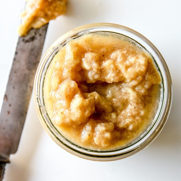 This is an overhead image of a knife with banana jam on the white counter next to a glass jar filled with banana jam. The jar sits on a white counter.