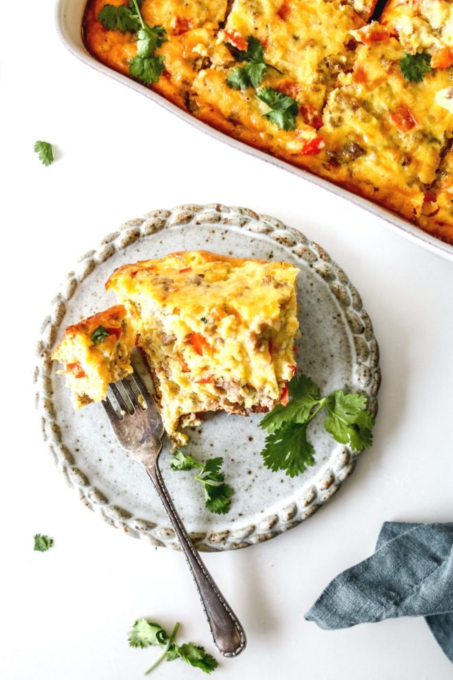 This is an overhead image of a grey speckled plate with a slice of egg casserole on it. A fork is cutting off and piercing a bite of the casserole. The piece is garnished with cilantro. The casserole dish with the casserole in it is in the top right of the image.
