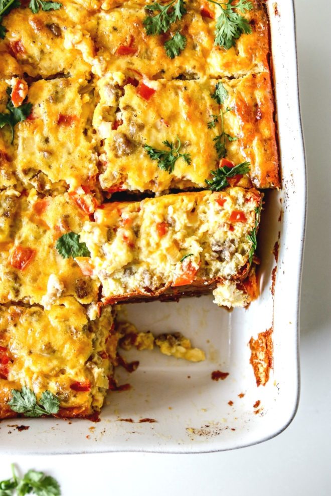 This is an overhead image of a breakfast casserole cut into 8 pieces in the casserole dish. One piece of the casserole is missing and one piece is turned on the side to show the peppers, onions, and sausage pieces in the casserole.