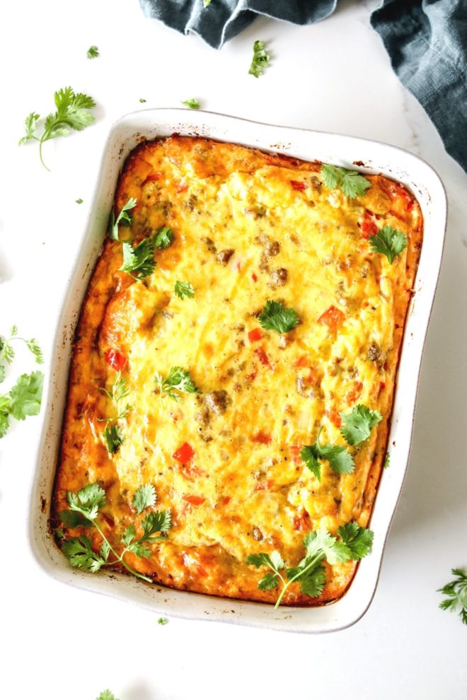 This is an overhead image of a white rectangle casserole dish with an egg, pepper, sausage, and onion breakfast casserole in it. The dish sits on a white counter with a blue tea towel on the top right corner. The casserole is garnished with cilantro.