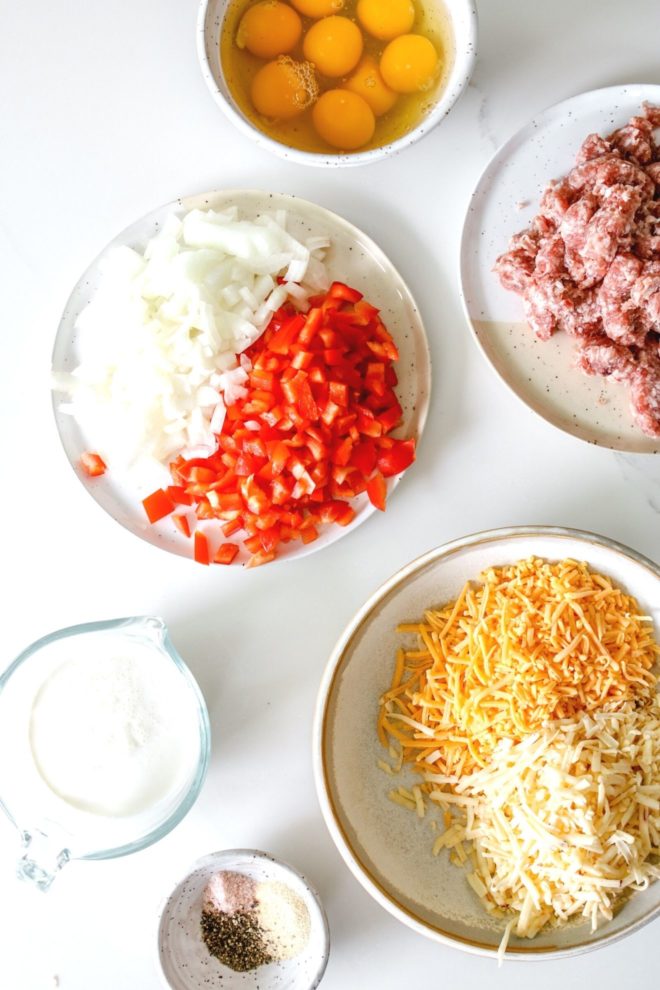 This is an overhead image of ingredients to make a breakfast casserole: eggs, onions, peppers, sausage, cream, shredded cheese, and spices on a white counter.