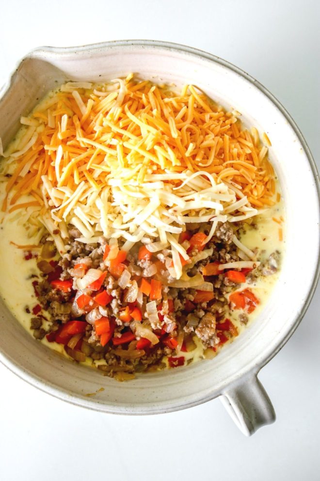 This is an overhead image of a large tan bowl with a spout and handle. Inside the bowl is whisked eggs, shredded cheese, and a pepper, onion and sausage mixture. The bowl sits on a white surface.