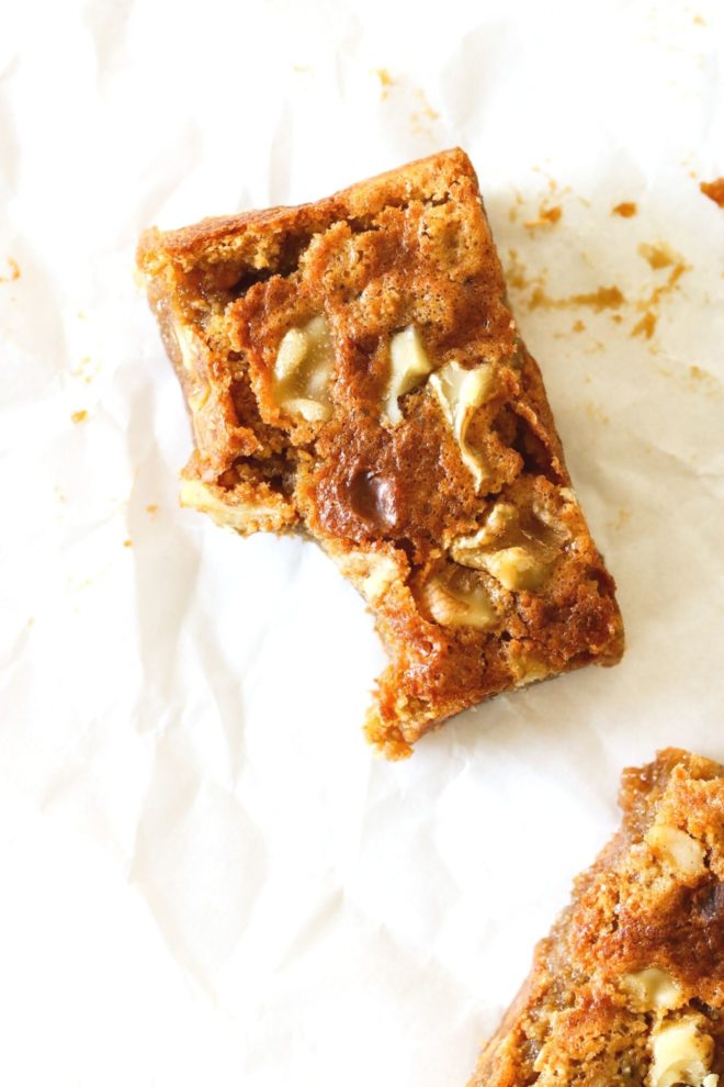 This is an overhead image of a butterscotch blondie with a bite taken out of it. The blondie sits on a white piece of parchment paper with another blondie in the bottom right corner of the image.