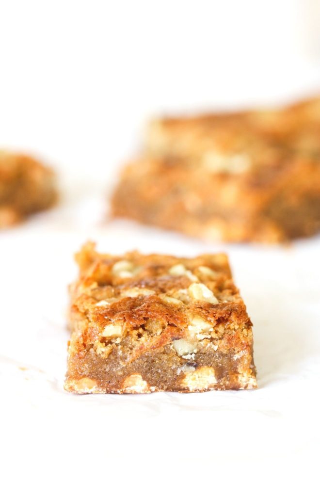 This is a side view of a butterscotch blondie with walnuts in it. The blondie sits on a white piece of parchment paper with more blondies blurred in the background.