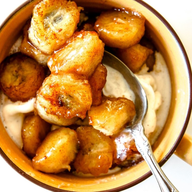 This is an overhead image of a yellow mug with vanilla ice cream and caramelized bananas. An antique spoon is scooping some of the bananas. The mug sits on a white counter.