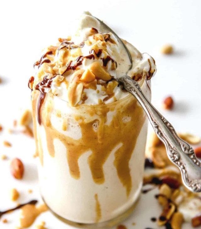 This is an overhead side view of a glass with a peanut butter milkshake in it. The milkshake is topped with whipped cream and drizzled with peanut butter and chocolate. An antique spoon is on the top of the glass. More drizzles and chopped peanuts are around the glass.