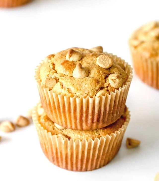 This is a side view of two peanut butter muffins stacked on top of each other. The muffins sit on a white counter with more muffins blurred in the background and peanut butter chips scattered around.