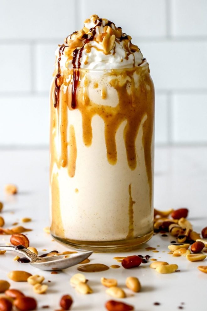 This is a side view of a glass with a peanut butter milkshake in it. The milkshake it topped with whipped cream and a peanut butter and chocolate drizzle. Peanuts are on top of the whipped cream and around the glass on the white counter.