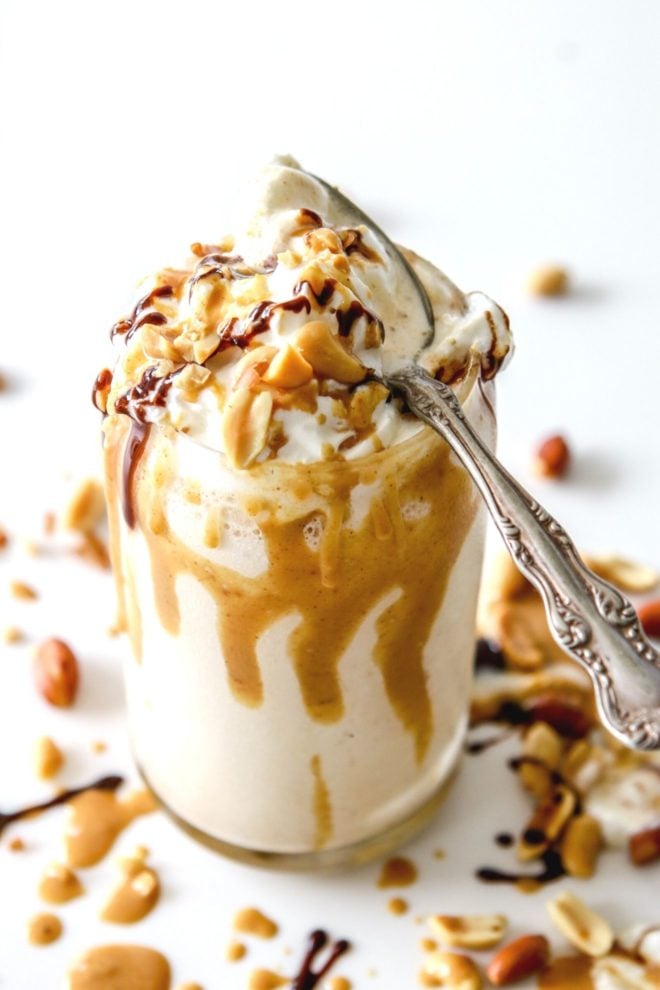 This is an overhead side view of a glass with a milkshake in it. The milkshake is topped with whipped cream and drizzled with peanut butter and chocolate. An antique spoon is on the top of the glass. More drizzles and chopped peanuts are around the glass.