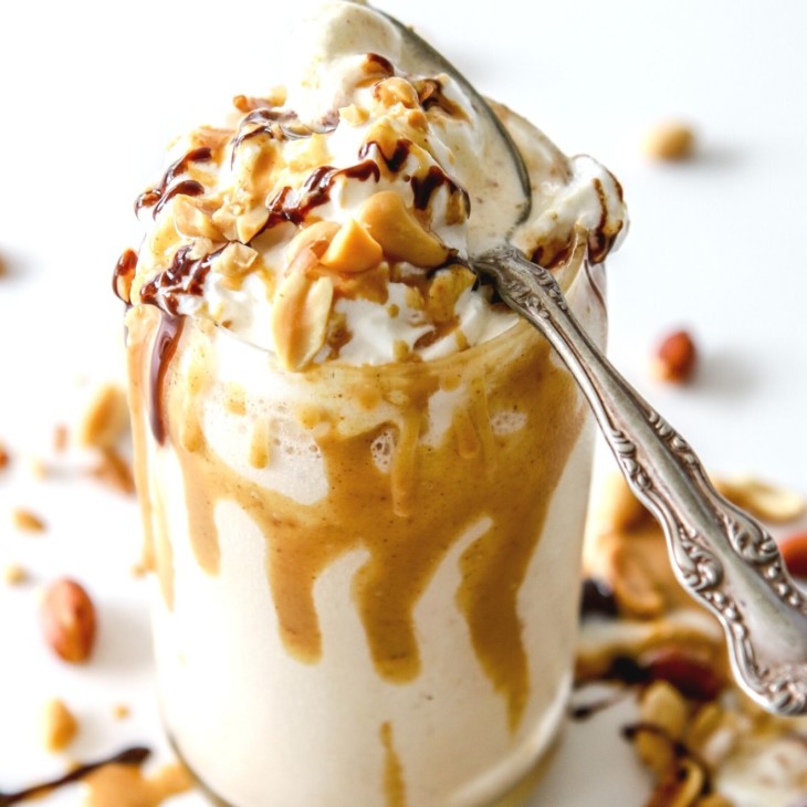 This is an overhead side view of a glass with a peanut butter milkshake in it. The milkshake is topped with whipped cream and drizzled with peanut butter and chocolate. An antique spoon is on the top of the glass. More drizzles and chopped peanuts are around the glass.