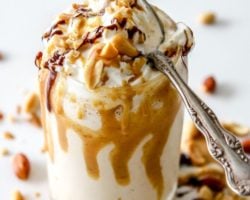 This is an overhead side view of a glass with a peanut butter milkshake in it. The milkshake is topped with whipped cream and drizzled with peanut butter and chocolate. An antique spoon is on the top of the glass. More drizzles and chopped peanuts are around the glass. Text overlay reads "peanut butter milkshake."