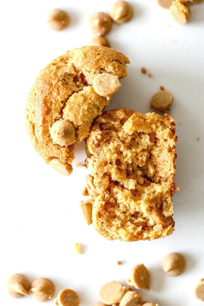 This is an overhead image of a muffin broke in half. Half of the muffin you can see the top of with peanut butter chips and the other half of the muffin is laying on its side so you can see the fluffy middle. The muffin lays on a white counter with peanut butter chips scattered around it.
