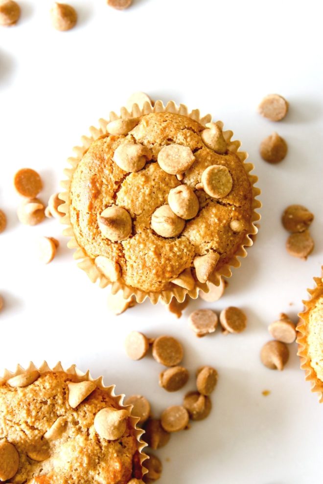This is an overhead image of a muffin with peanut butter chips on top. The muffin sits on a white counter with more muffins to the bottom left and right of the image. Peanut butter chips are scattered on the white counter.