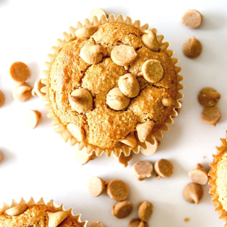 This is an overhead image of a peanut butter muffin with peanut butter chips on top. The muffin sits on a white counter with more muffins to the bottom left and right of the image. Peanut butter chips are scattered on the white counter.